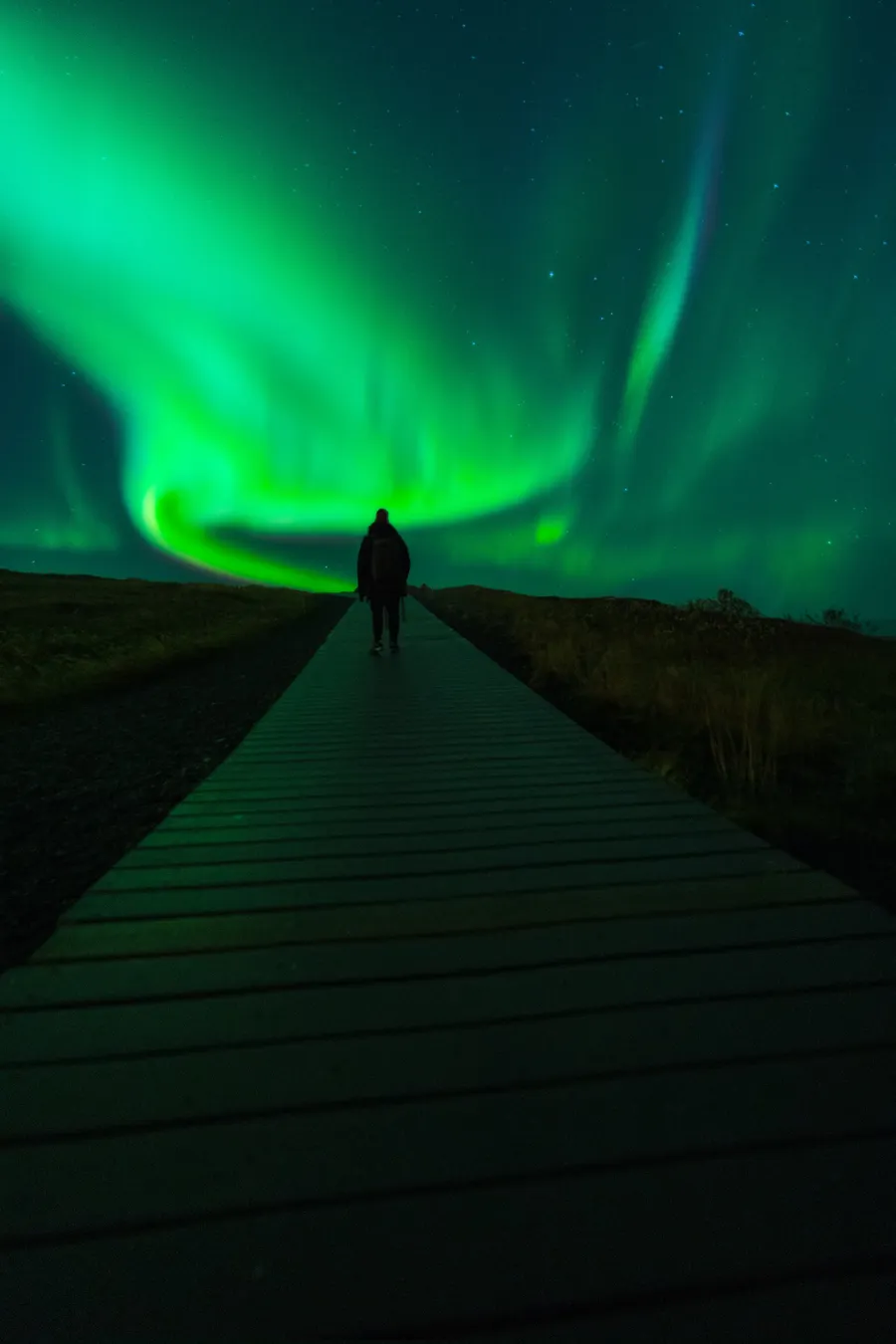 Una persona caminando por una pasarela de madera hacia una aurora verde en espiral